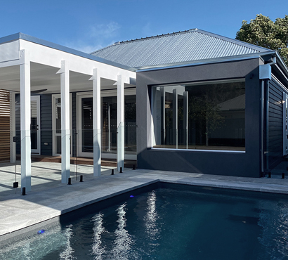 Modern extension to the rear of a traditional heritage home by local Williamstown architect Dig Design featuring a large window seat overlooking a swimming pool surrounded with bluestone paving. A roofed white pergola covering an outdoor entertainment area is next to the pool.