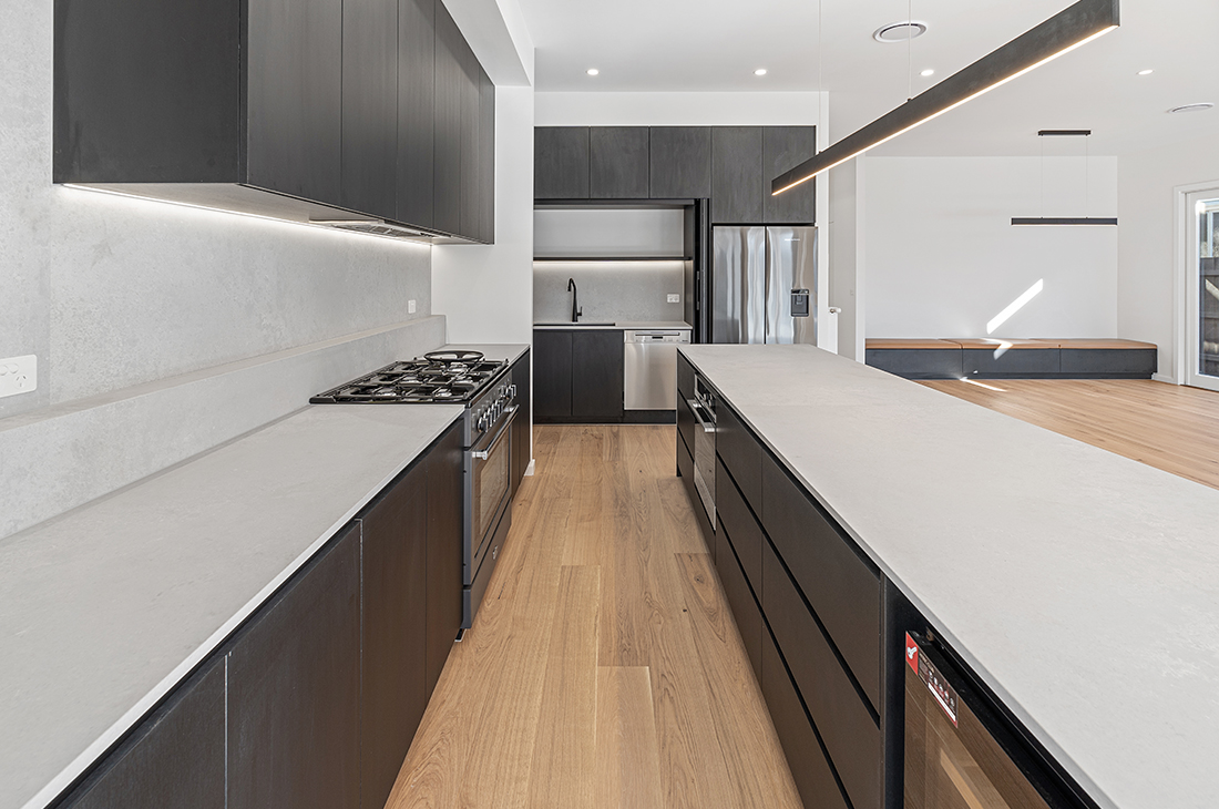 Large modern kitchen featuring black cabinets, grey concrete coloured stone benchtops and full height splashback. The pocket door cabinets in the background are open, exposing the sink black tapware and dishwasher. The large black Falcon stove is on the right. A large black lineal pendant is used over the island bench and undercabinet LED strips illuminate benchtops along the walls. Light coloured engineered oak flooring is used. The tan leather upholstered bench seat to the dining area can be seen in the background to the right.