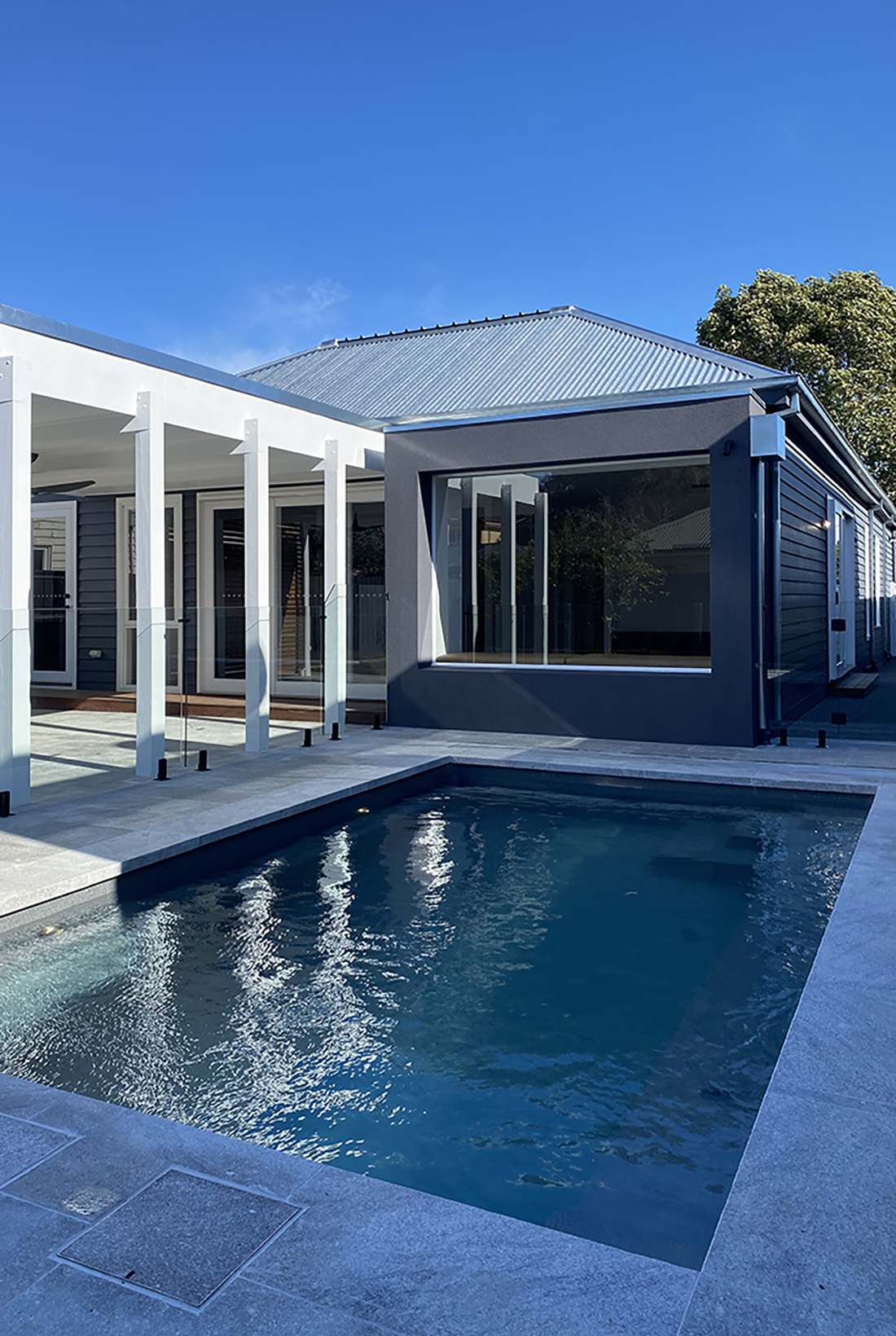 Modern extension to the rear of a traditional heritage home by local Williamstown architect Dig Design featuring a large window seat overlooking a swimming pool surrounded with bluestone paving. A roofed white pergola covering an outdoor entertainment area is next to the pool.