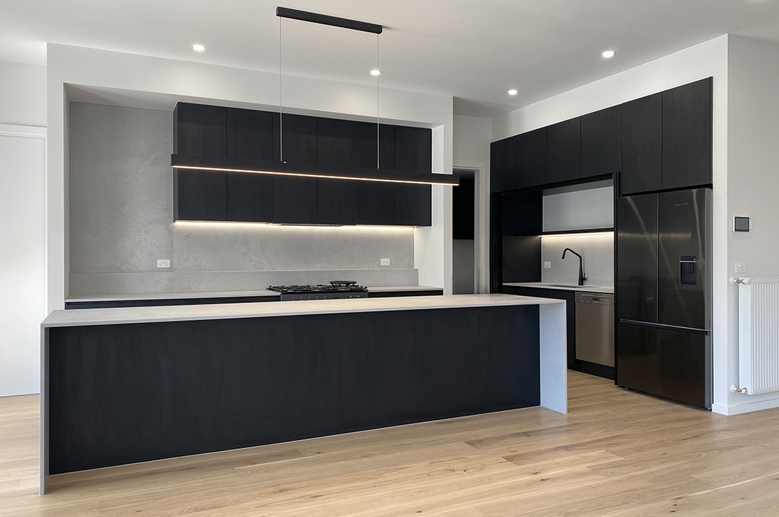 Modern kitchen by Williamstown interior designer Dig Design features black cabinets, grey concrete coloured stone benchtops and full height splashback. The pocket door cabinets on the right are open, exposing the sink black tapware and dishwasher. The space is filled with natural light from the large glass doors out of shot on the left. A large black lineal pendant is used over the island bench and undercabinet LED strips illuminate benchtops along the walls, there are also recessed downlights illuminating the space. Light coloured engineered oak flooring is used.