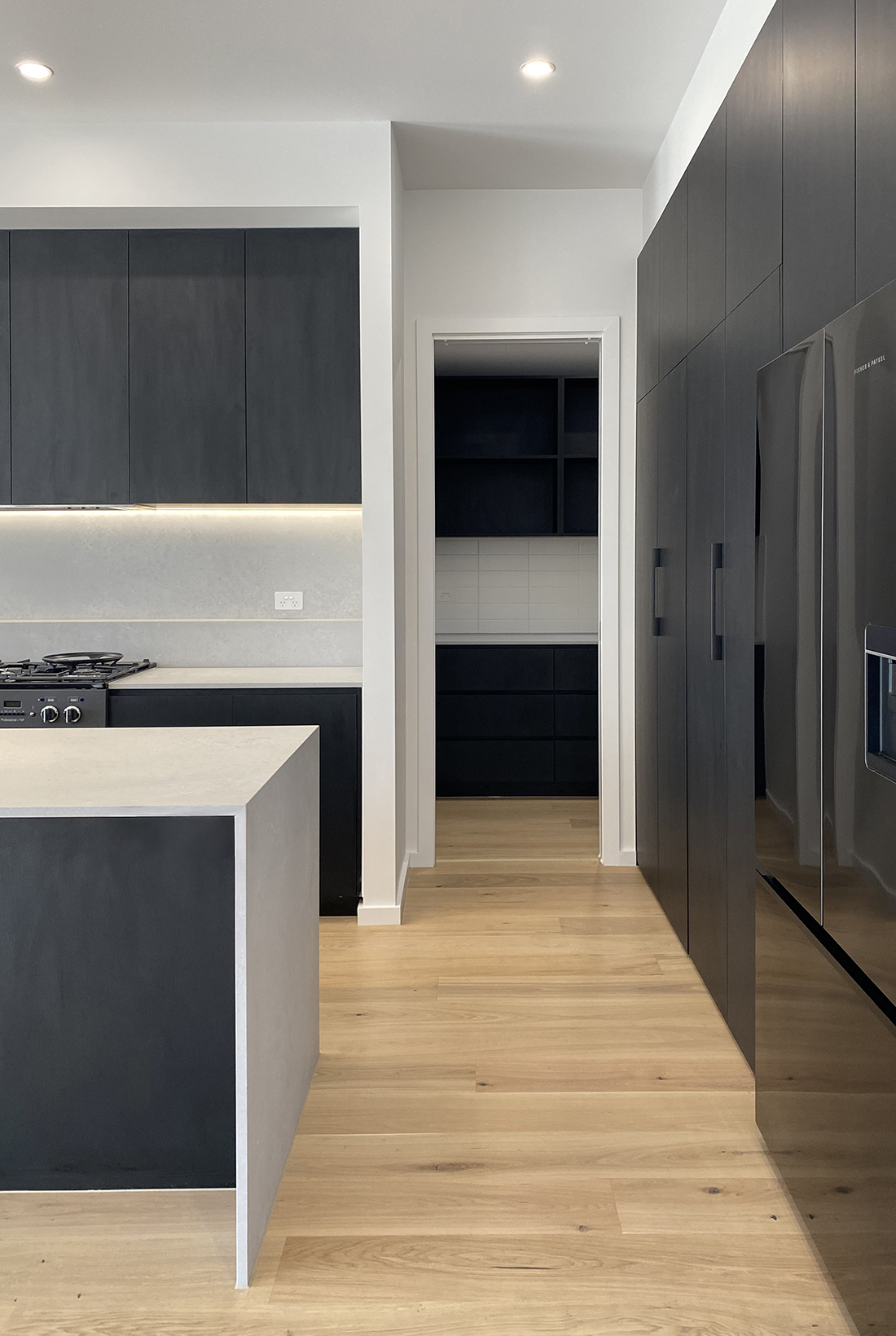 Modern kitchen with black cabinets and concrete coloured stone benchtops and splashback. A hint of the Falcon stove can be seen, as can the black cabinetry in the pantry beyond. Pocket door cabinets hide the sink and dishwasher on the right and the dark stainless steel fridge can also be seen. Light coloured engineered oak floorboards are used throughout. The space is illuminated with natural light form large glass doors to the left, recessed downlights above and an undercabinet LED light strip above the kitchen bench.