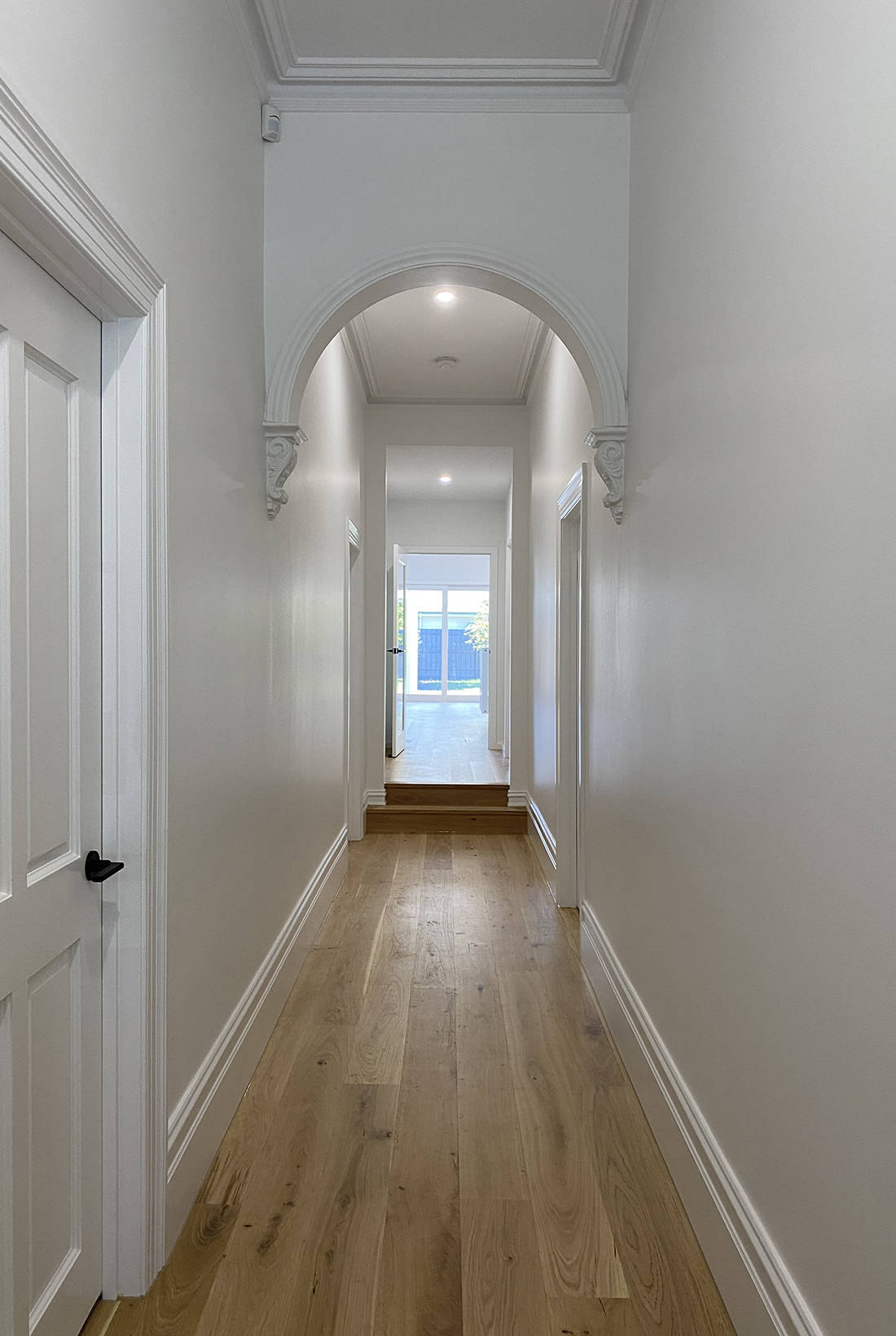 White Victorian hallway with high ceilings, traditional skirts, cornices and archways. Light coloured timber floor with white walls. At the end of the hallway, the light shines in through the glass doors to the outdoor entertainment area.