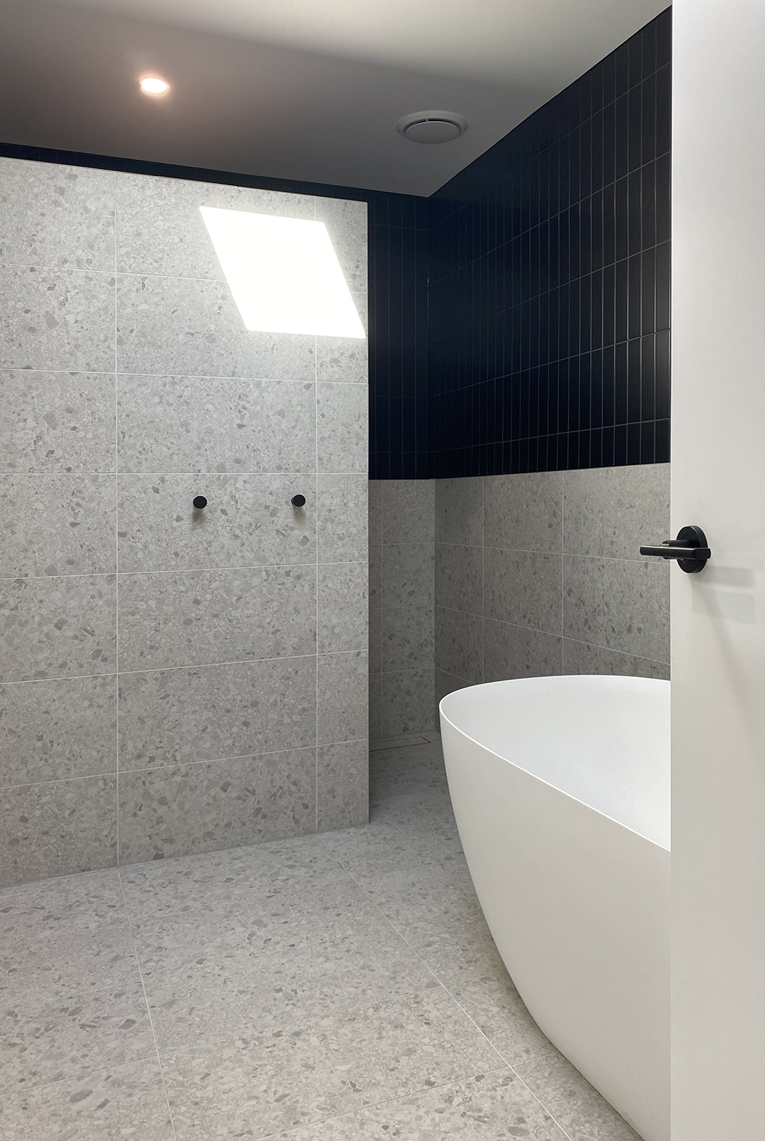 Modern bathroom featuring a freestanding white egg shaped bathtub. The space is filled with natural light from a skylight above. A reduced height partition wall hides the shower and toilet and is tiled with large format grey terrazzo stone look tiles, with two black robe hooks. The walls are tiled with a black subway tile on the top half of the wall and the grey terrazzo stone look tiles below and across the floor. A hint of the bathroom door can be seen with its accent black door handle.