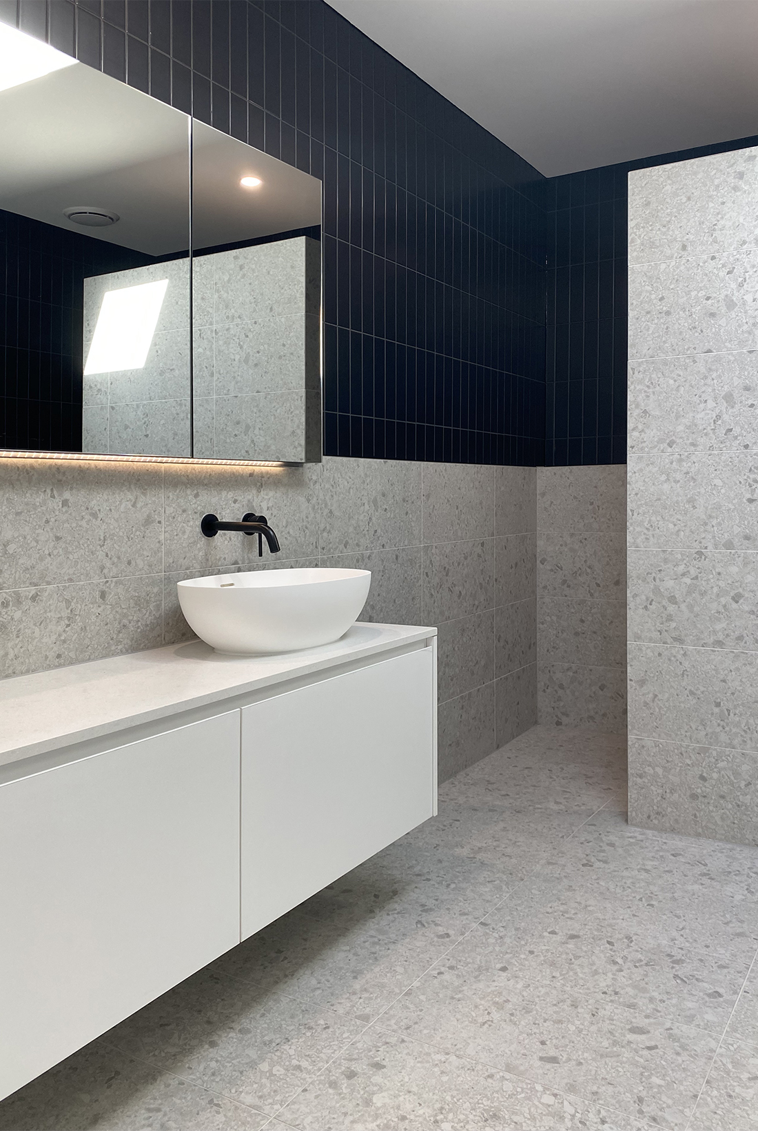 Modern bathroom by Williamstown interior designer Dig Design featuring a floating white vanity with stone top, an above counter sink and black tapware. Above the vanity is a large mirrored shaving cabinet. The walls have a combination of black subway tiles and grey terrazzo stone look tiles, the floor has the same grey terrazzo stone look tiles as does the reduced height partition wall. A skylight illuminates the space.