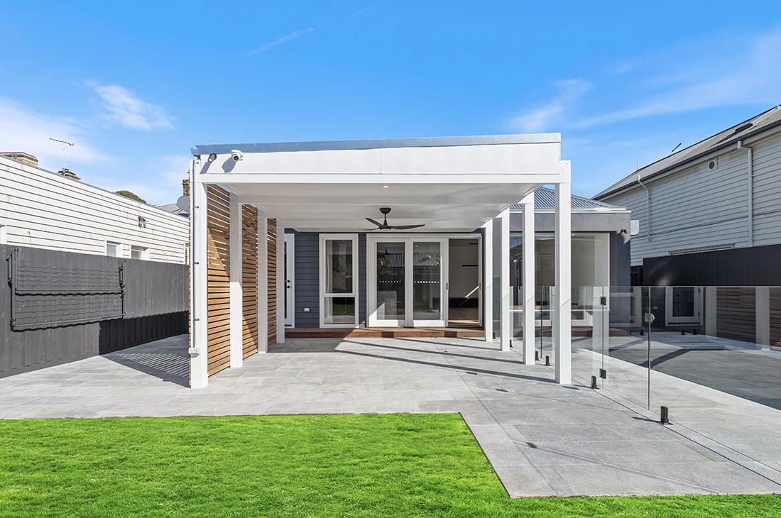 Roofed white pergola coming off a modern extension to the rear of a traditional heritage home in Williamstown with bluestone paving and a timber batten screen to one side. The pergola creates a light and bright outdoor entertainment area and has a ceiling fan. Next to the pergola is a glass pool fence and a swimming pool. The sky is blue and there is a patch of green grass in the foreground.